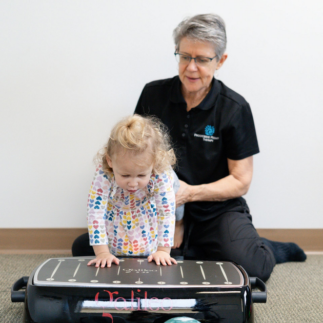 Woman assisting toddler on a black therapy barrel in a clinical room