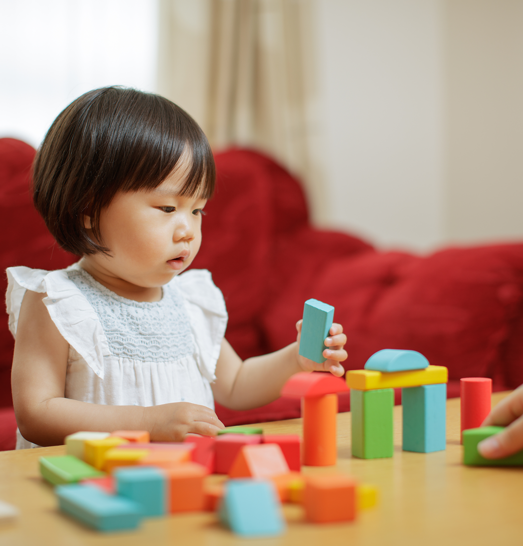 Child playing with colorful blocks at a table in a living room, reaching for a blue piece.