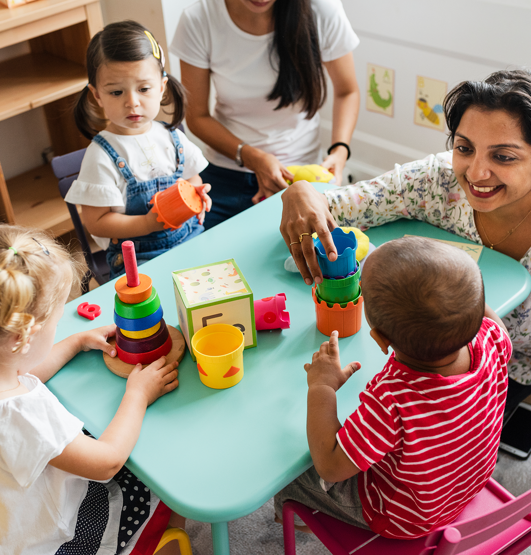 Children playing with colorful toys at a table while an adult supervises in a bright room