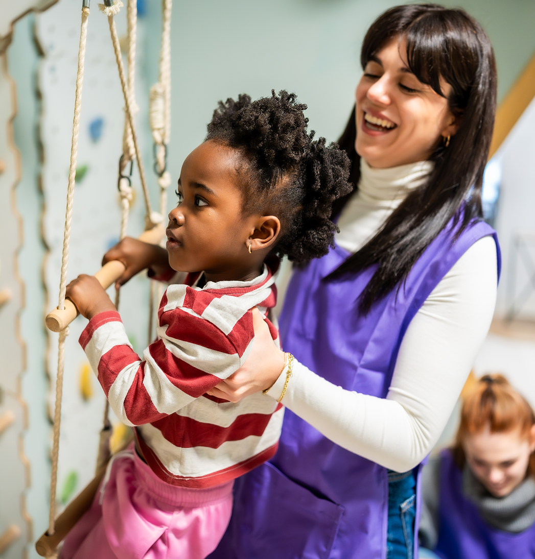 Adult helping a child on a rope swing indoors, both smiling