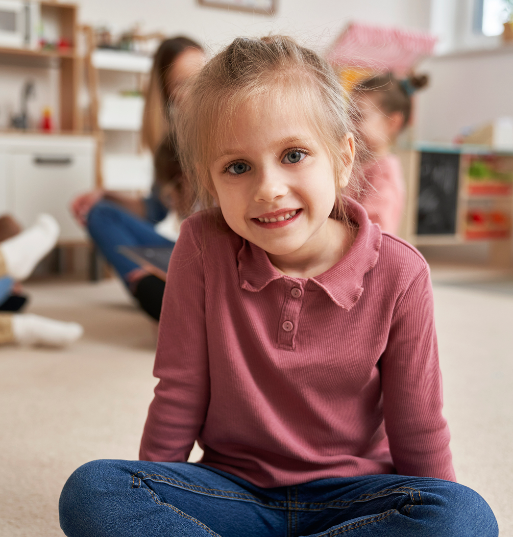 Smiling child sitting cross-legged on the floor in a playroom, wearing a pink shirt and jeans.
