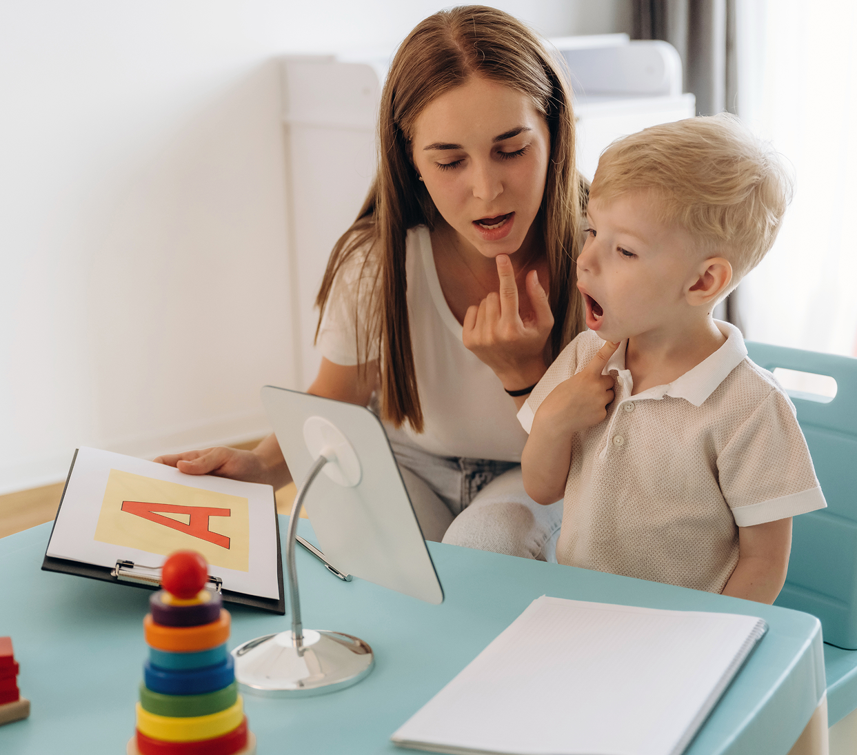Woman helping a young child at a desk with learning materials and a tablet in a bright room