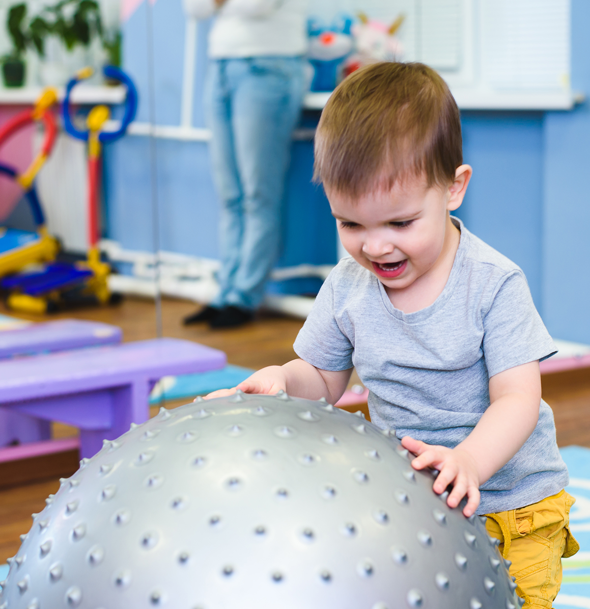Toddler smiling while playing with a large gray exercise ball in a colorful playroom