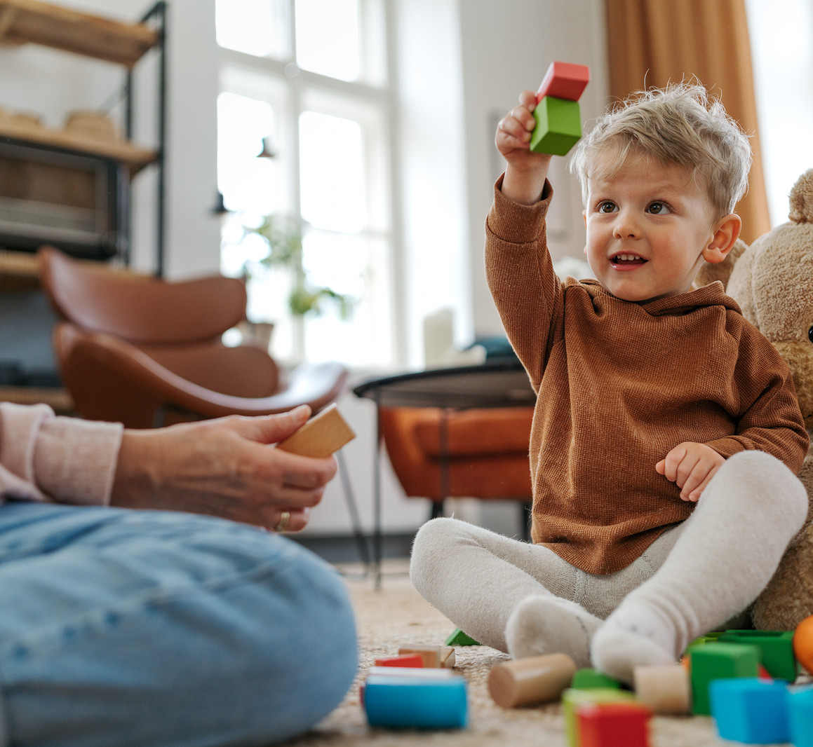 Toddler playing with colorful blocks in a cozy living room, holding up a green and red toy cube