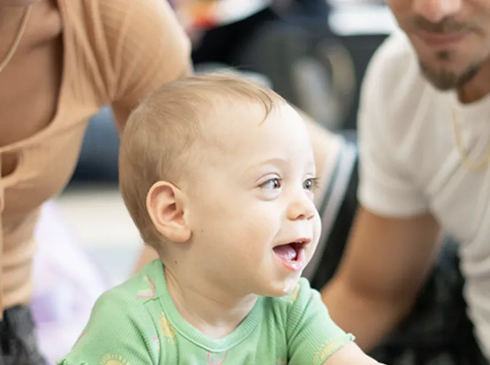 Smiling baby in green shirt, with adults nearby in a bright indoor setting