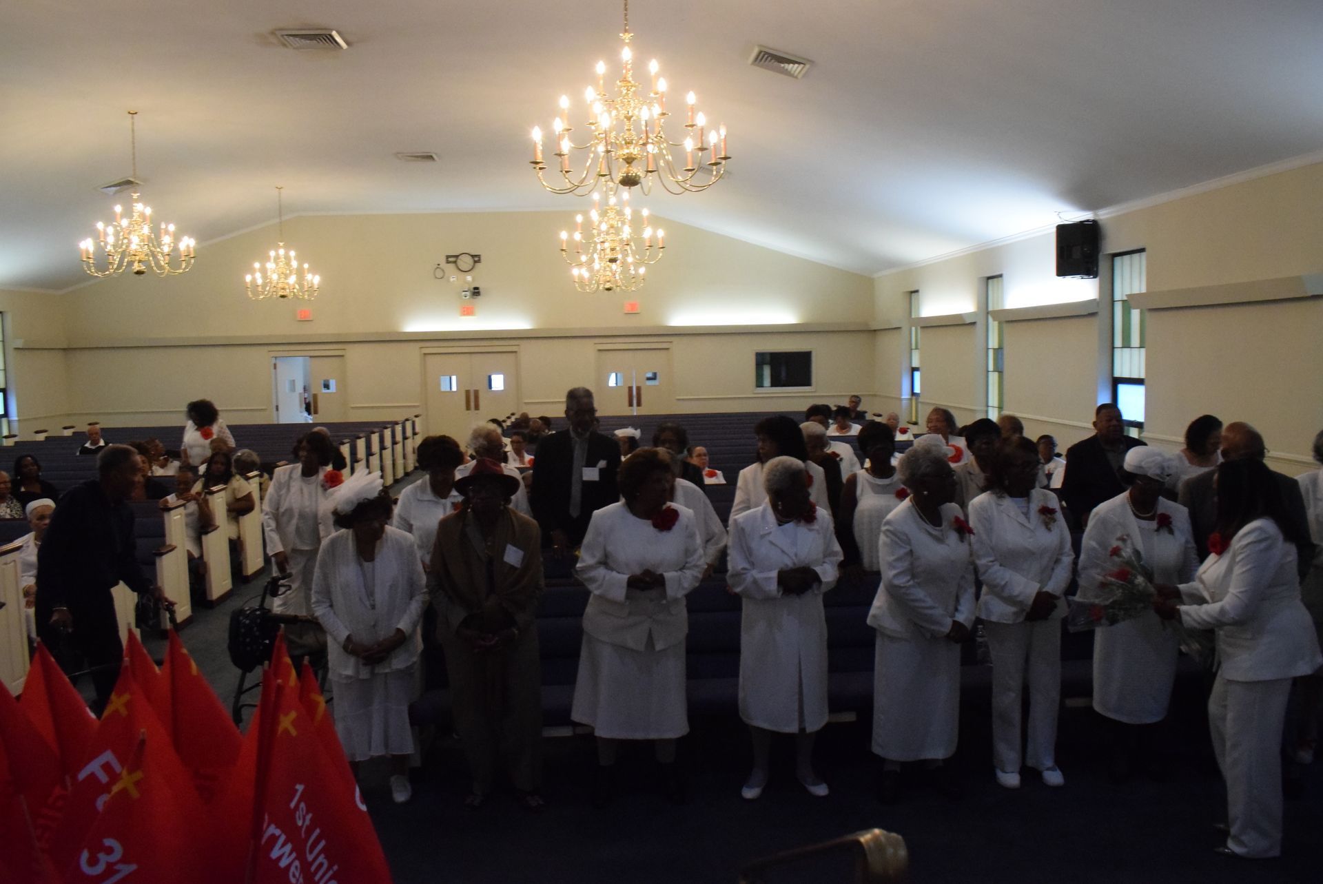 Women in white uniforms standing in a church sanctuary with chandeliers and red flags.