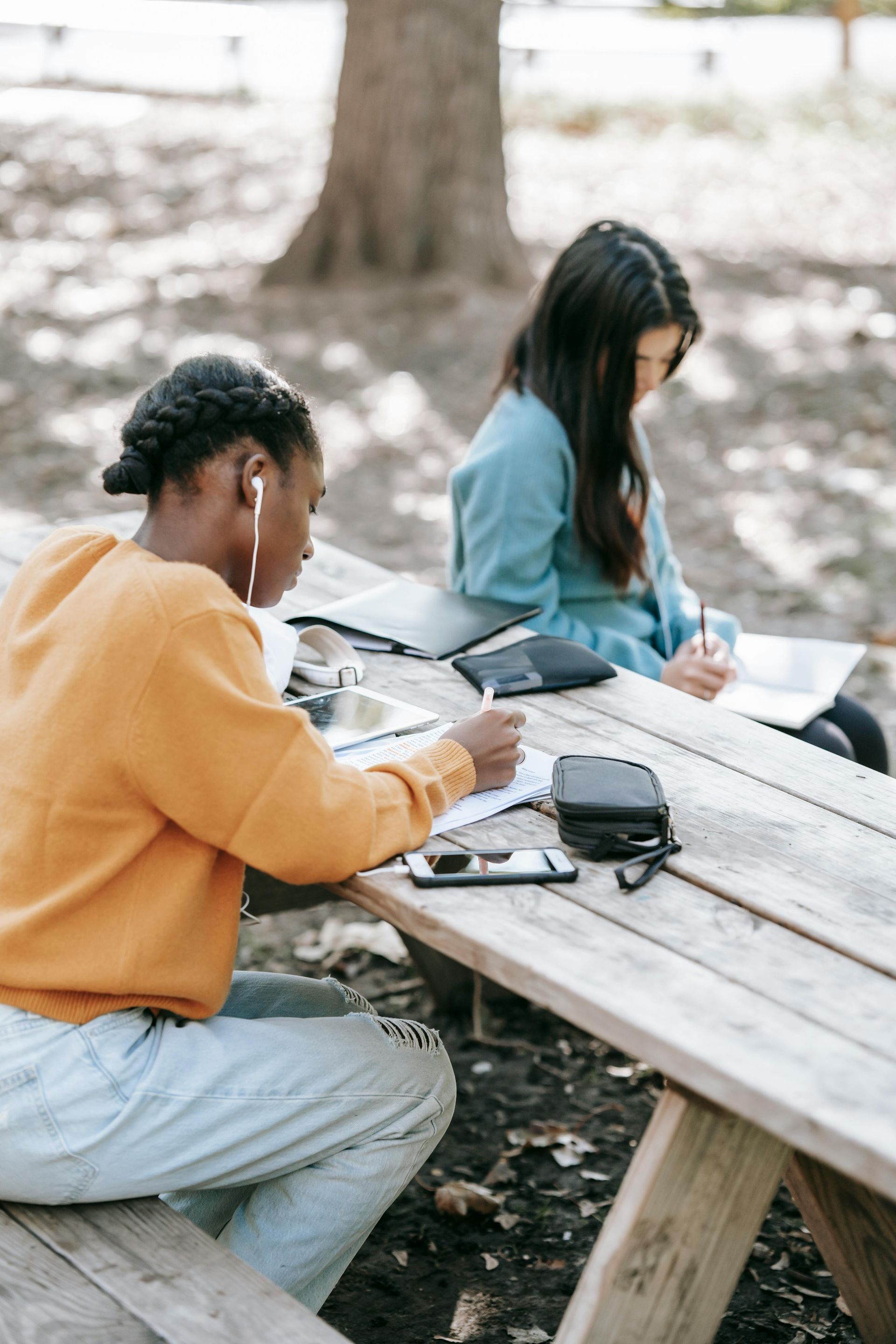 Two people sitting at a picnic table outdoors, looking down at notebooks and a phone.