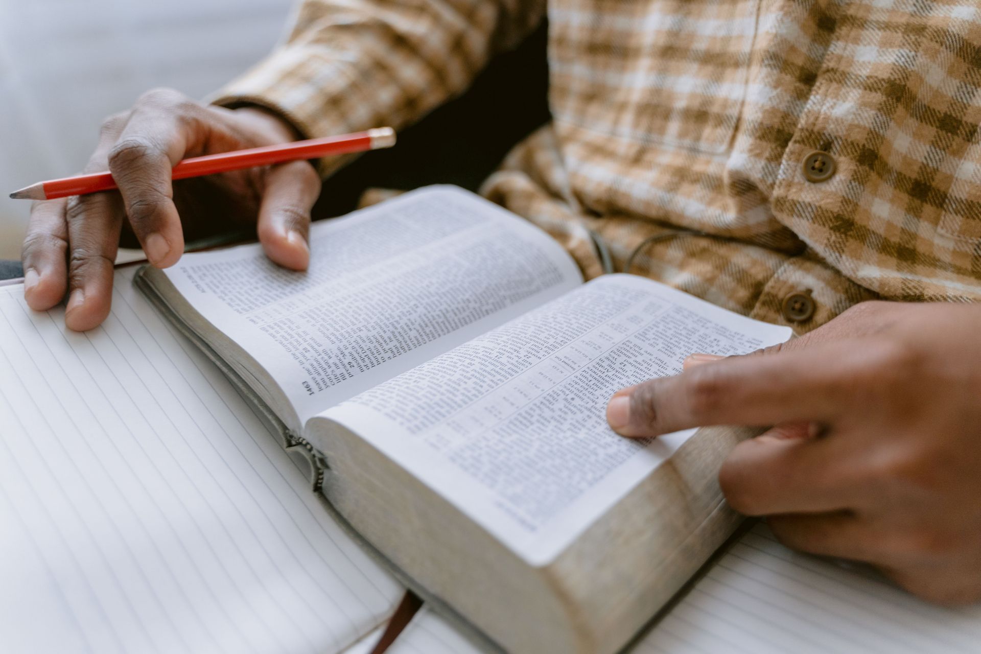 Hands holding an open bible and red pencil on a table, reading and taking notes