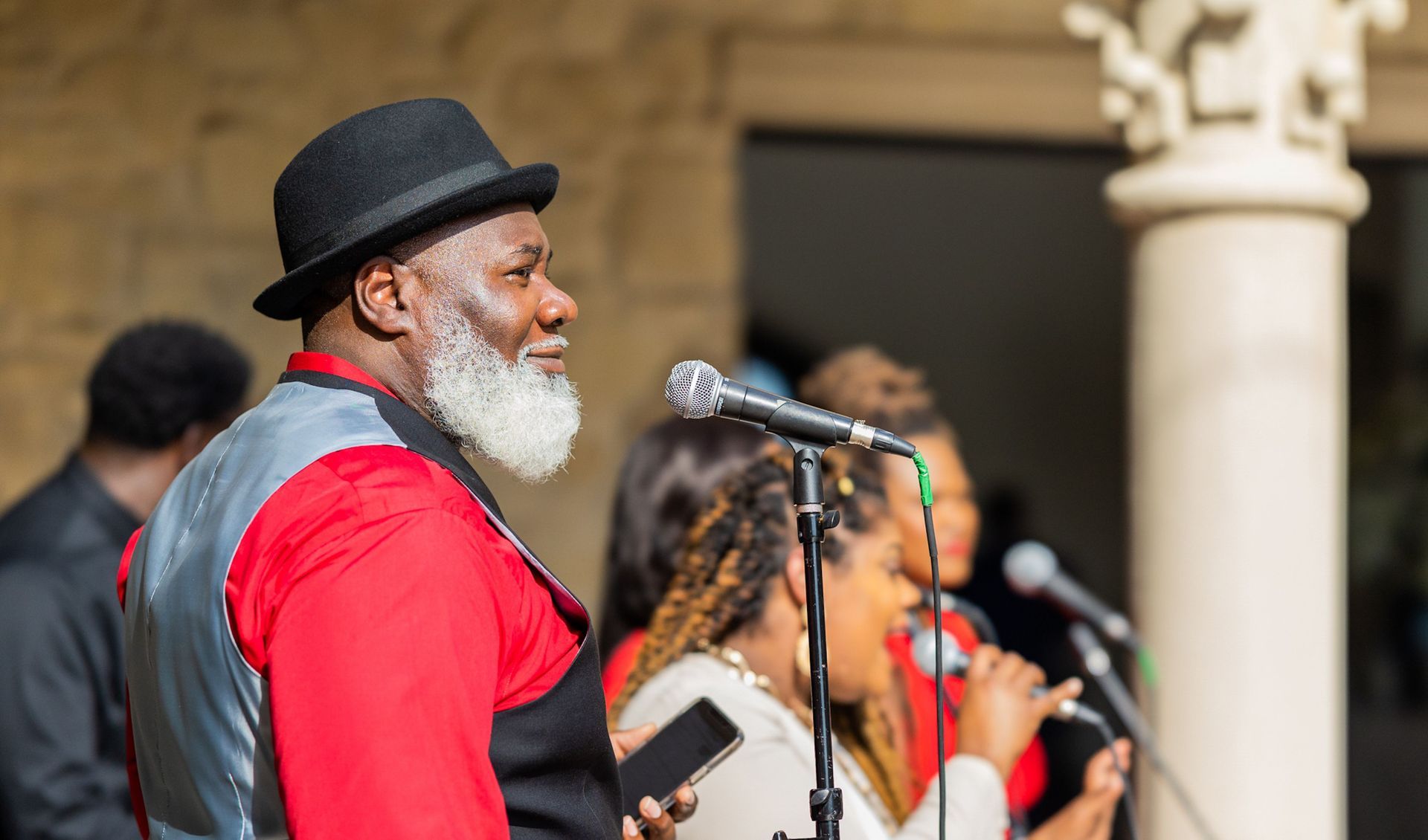 Musicians performing outdoors, with a man in a black hat and red shirt singing into a microphone.