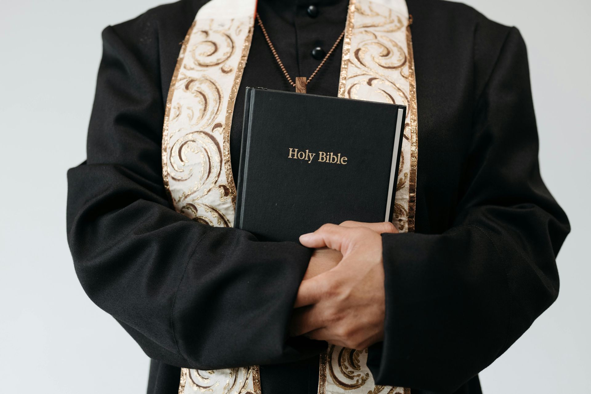 Clergyman in a black robe with an embroidered stole holding a black bible against their chest
