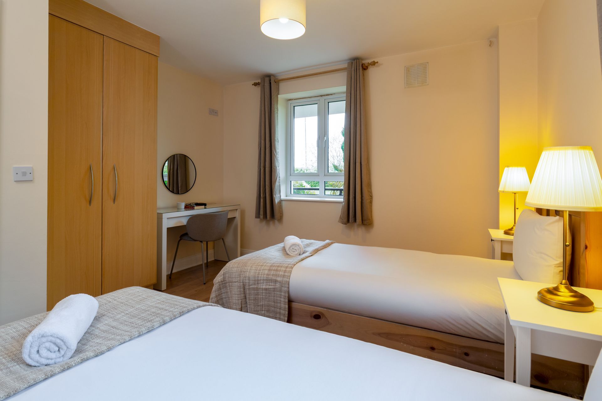 Two twin beds in a bedroom with a window, desk, wardrobe, and lamps.