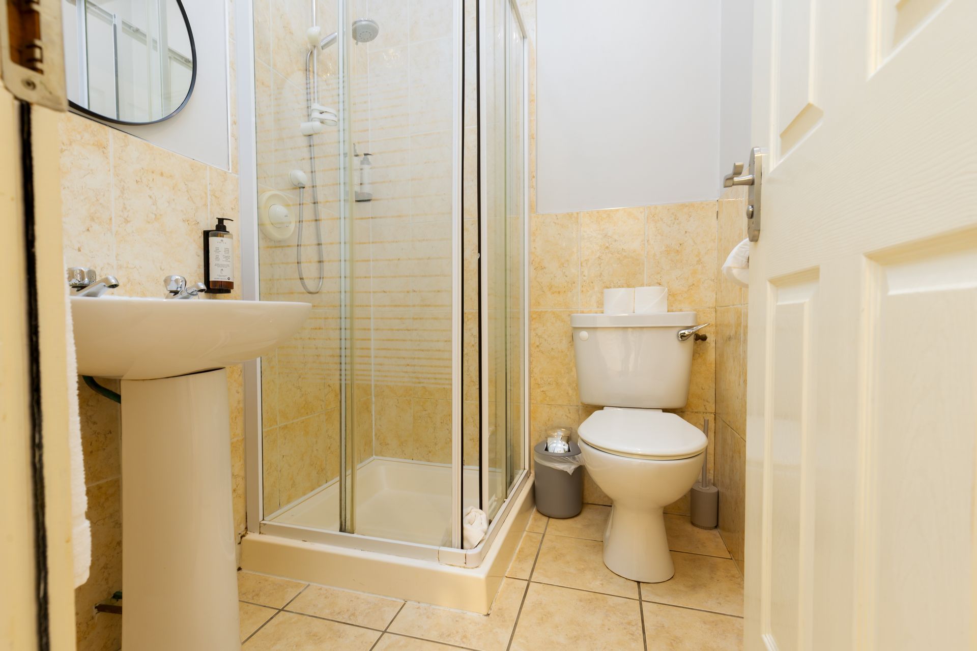 Bathroom with a pedestal sink, shower stall, and toilet. Beige walls and tiled floor.
