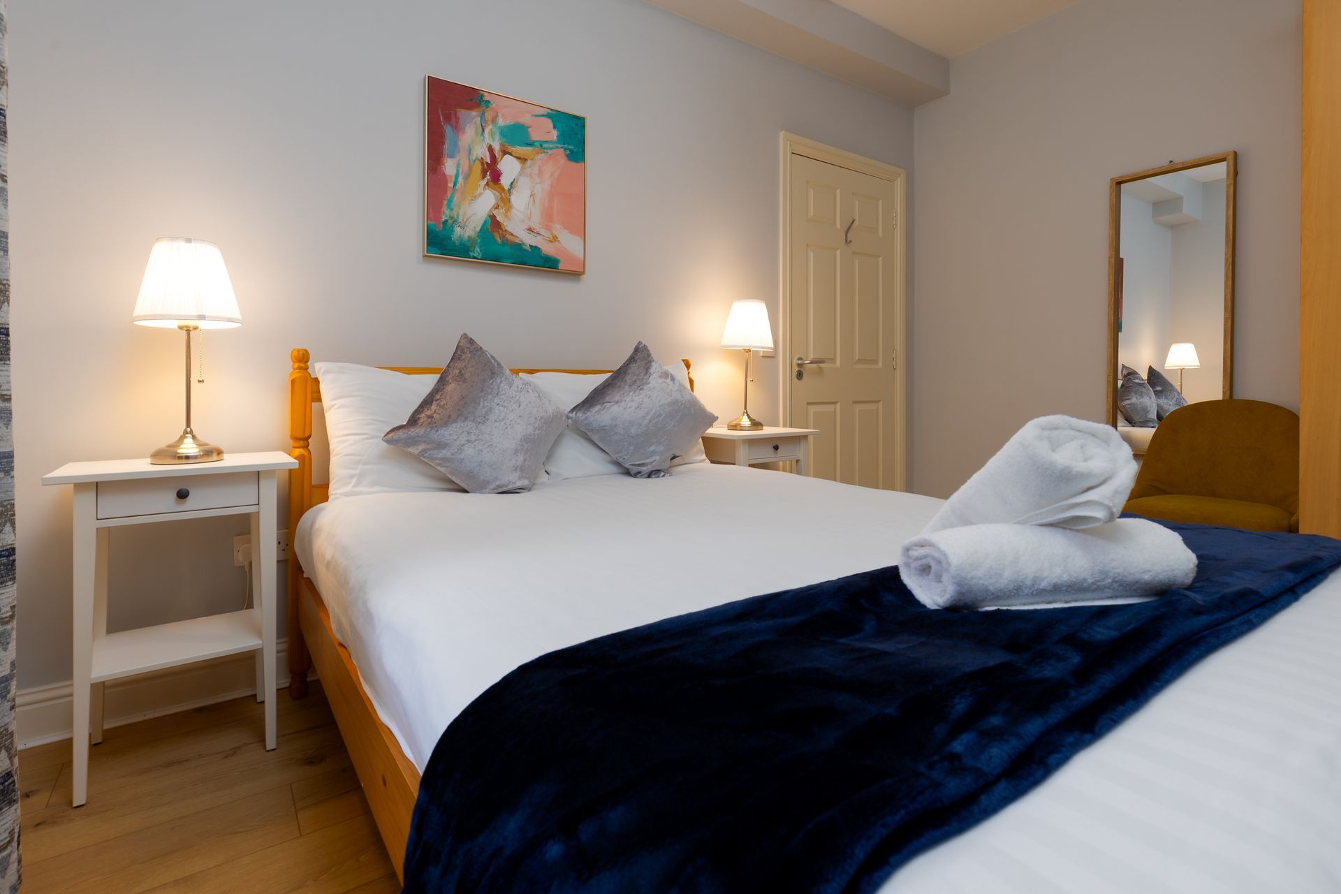 Bedroom with double bed, white linens, two lamps, and artwork. A towel is rolled on the bed.