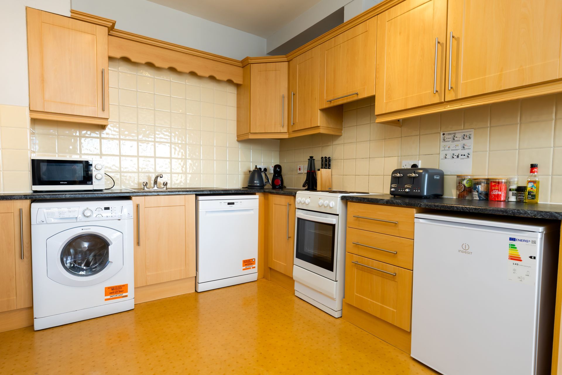 Kitchen with wooden cabinets, appliances including washer, microwave, and refrigerator. Yellow floor and tiled backsplash.