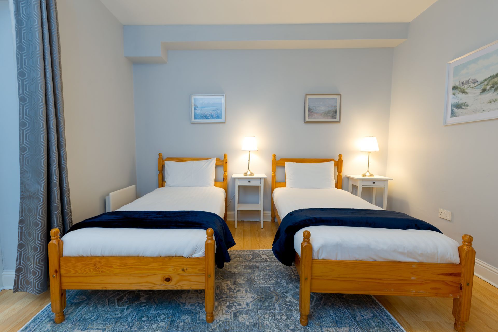 Two twin beds in a room with white bedding, wooden frames, and nightstands. Blue accents and artwork.