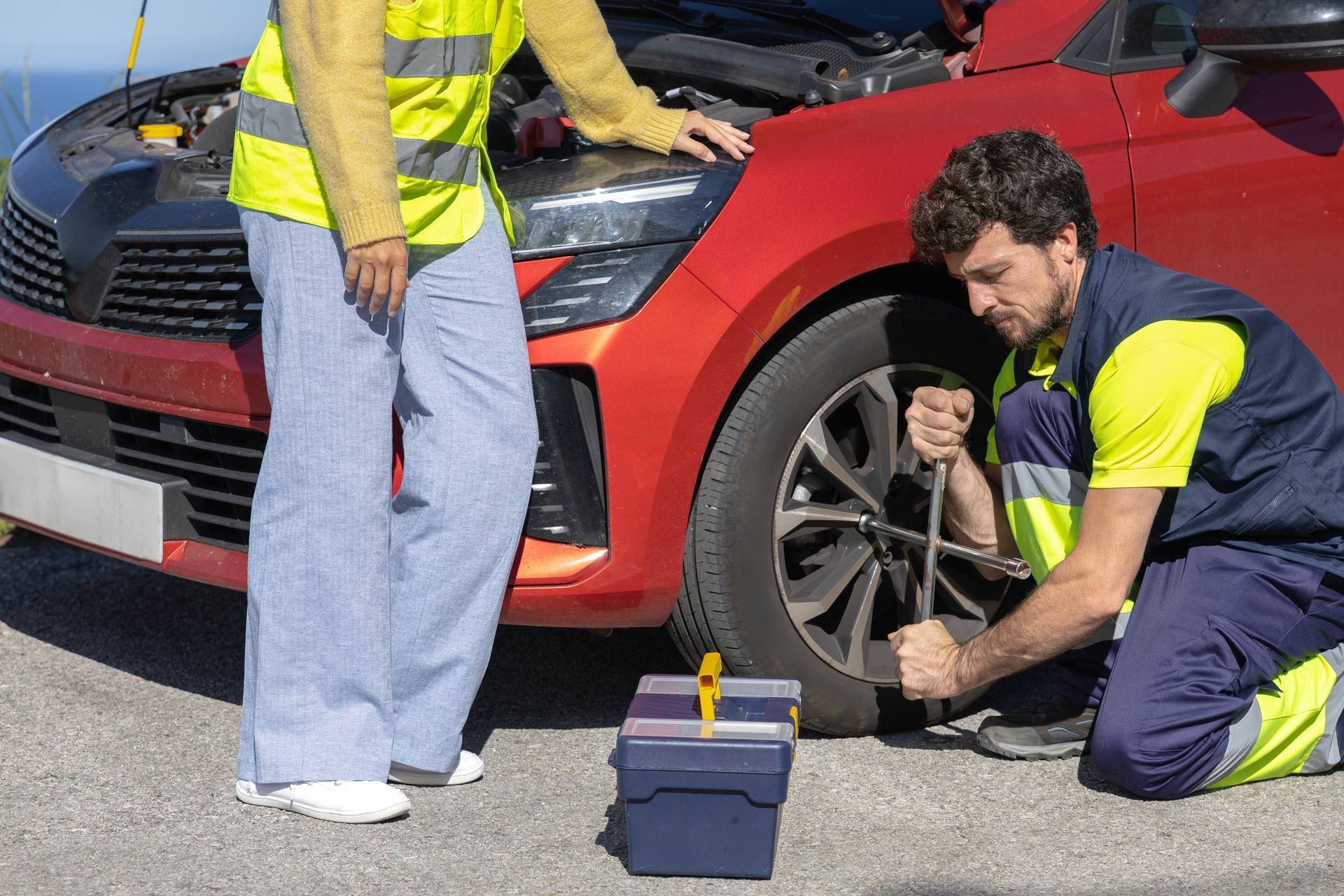 Mechanic changing a car tire beside a red vehicle on the roadside