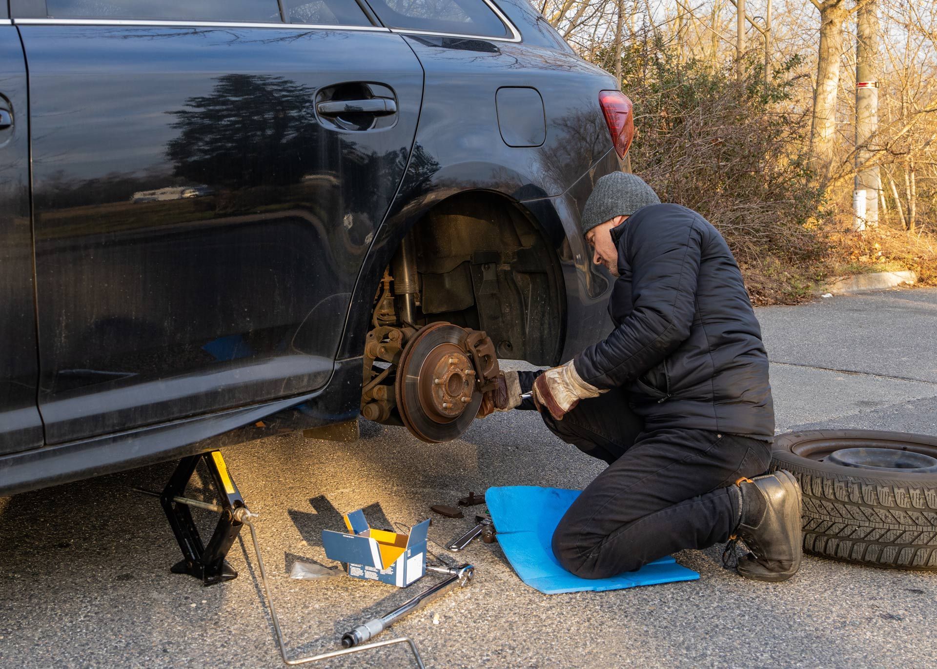 Mechanic kneeling beside black SUV, changing a flat rear wheel on the roadside.