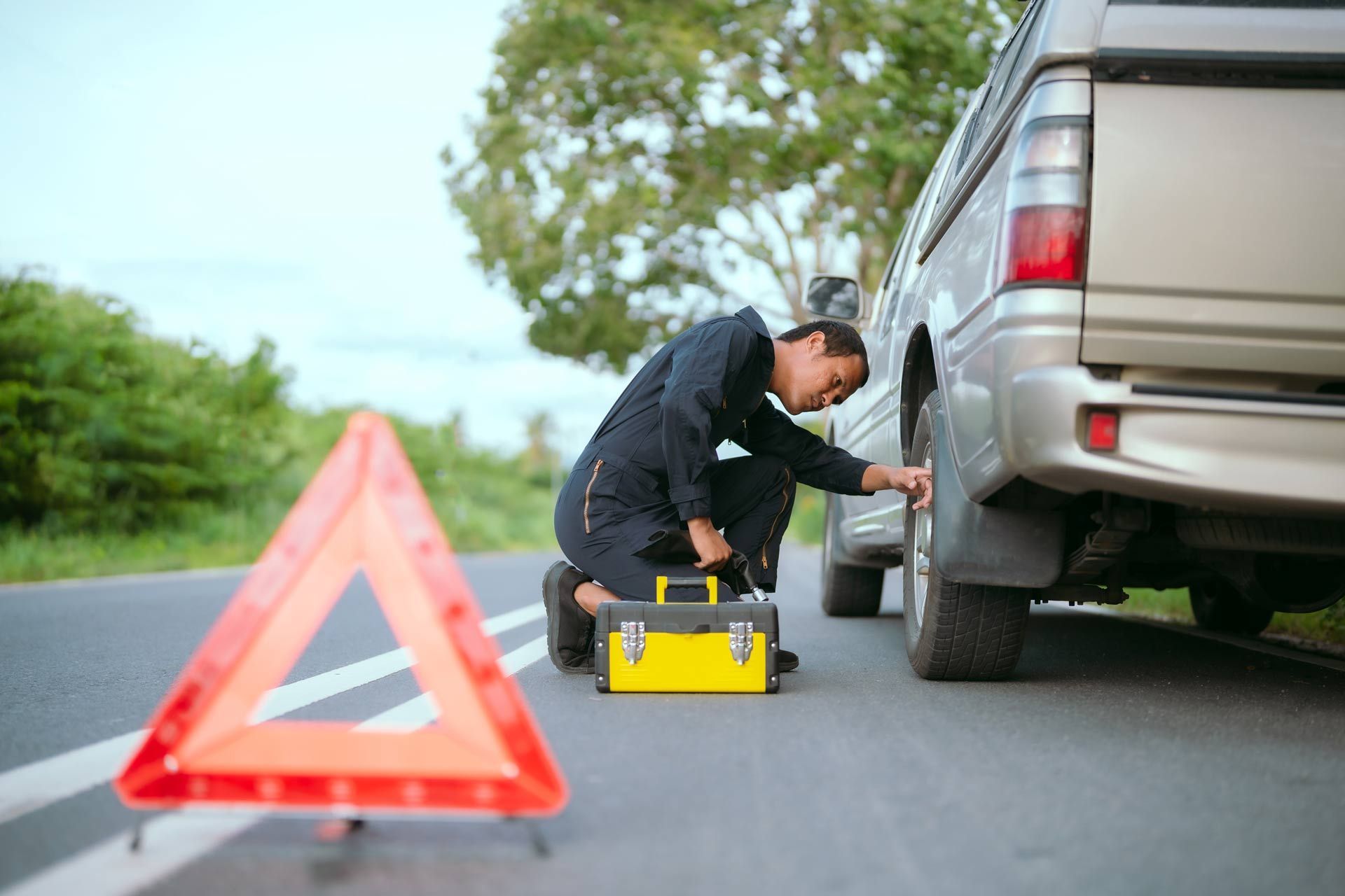 Driver crouching by a silver SUV with a warning triangle on the roadside, inspecting the rear wheel