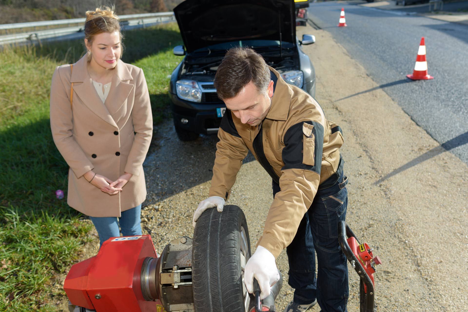 Two people inspect a car tire near a roadside with traffic cones and an open hood.