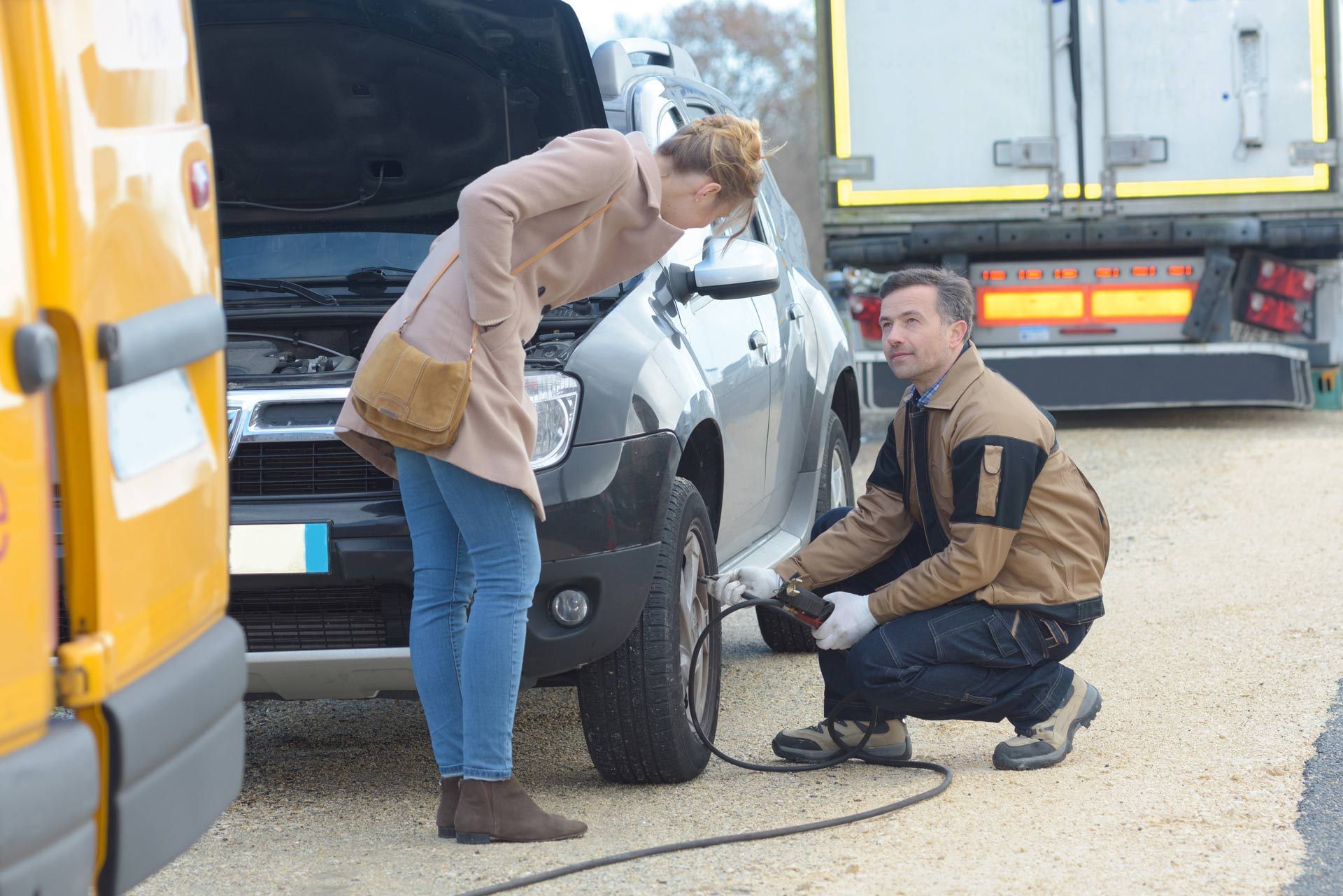 Person crouching by a silver car’s front tire while another leans in, on a roadside near a truck.