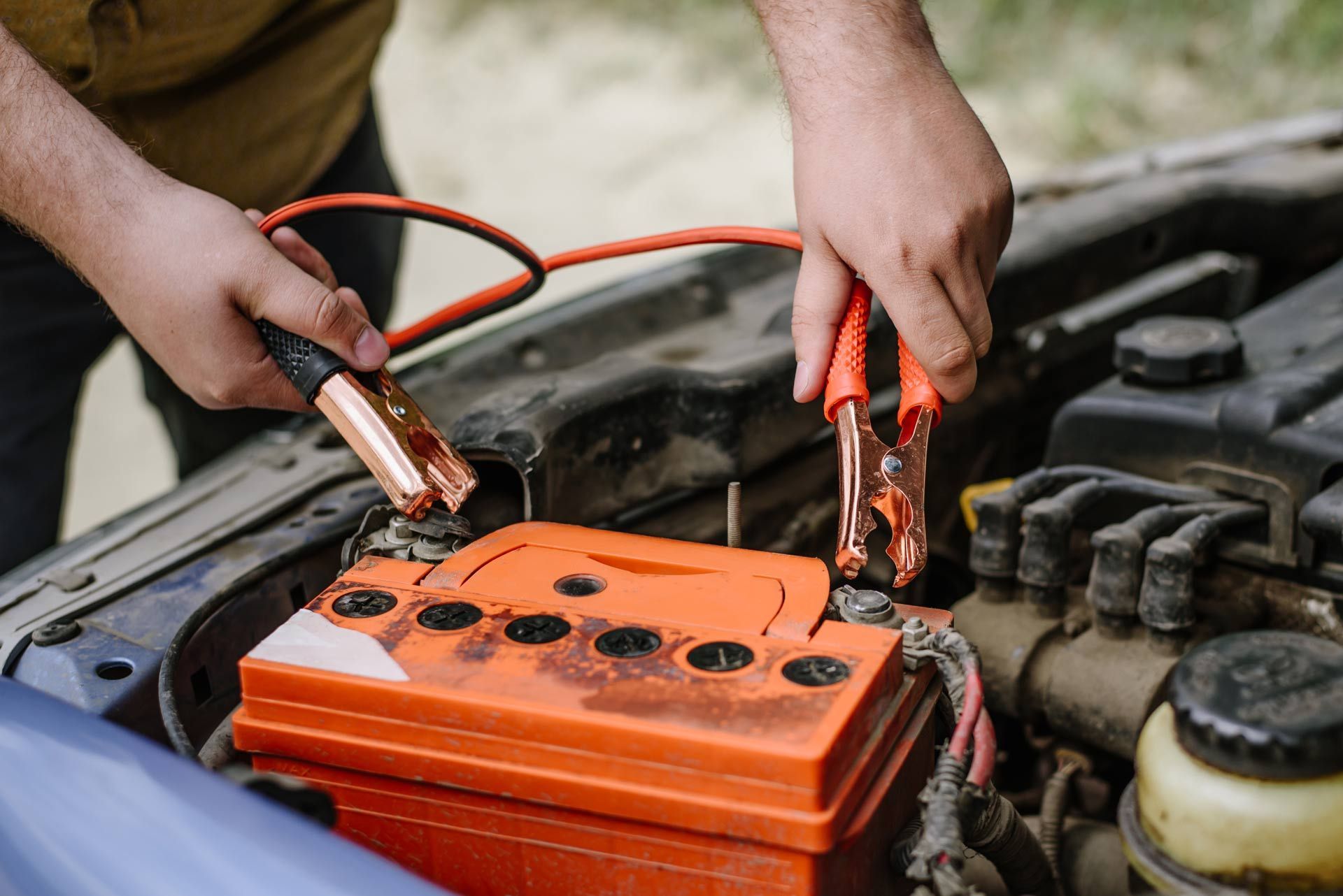 Hands using jumper cables to jump-start an orange car battery in an engine bay