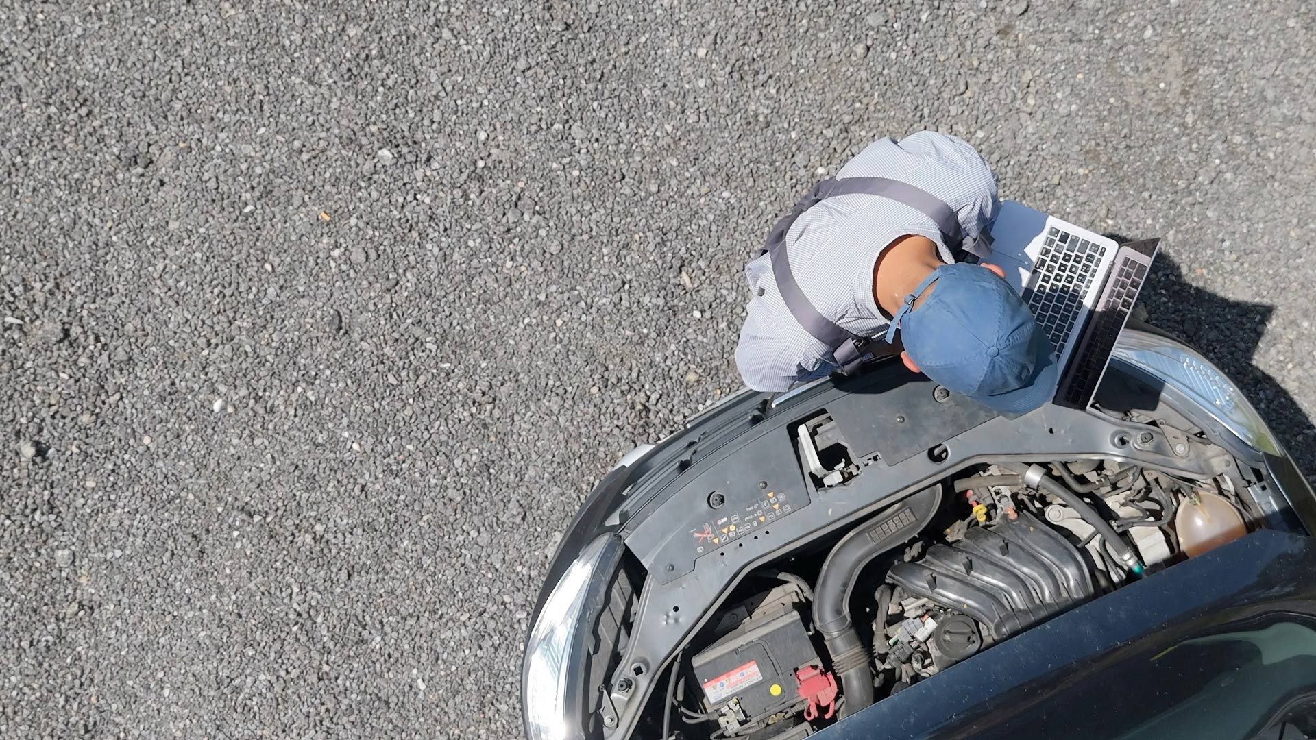 Person in blue cap working on a car engine beside the open hood on a gravel surface.