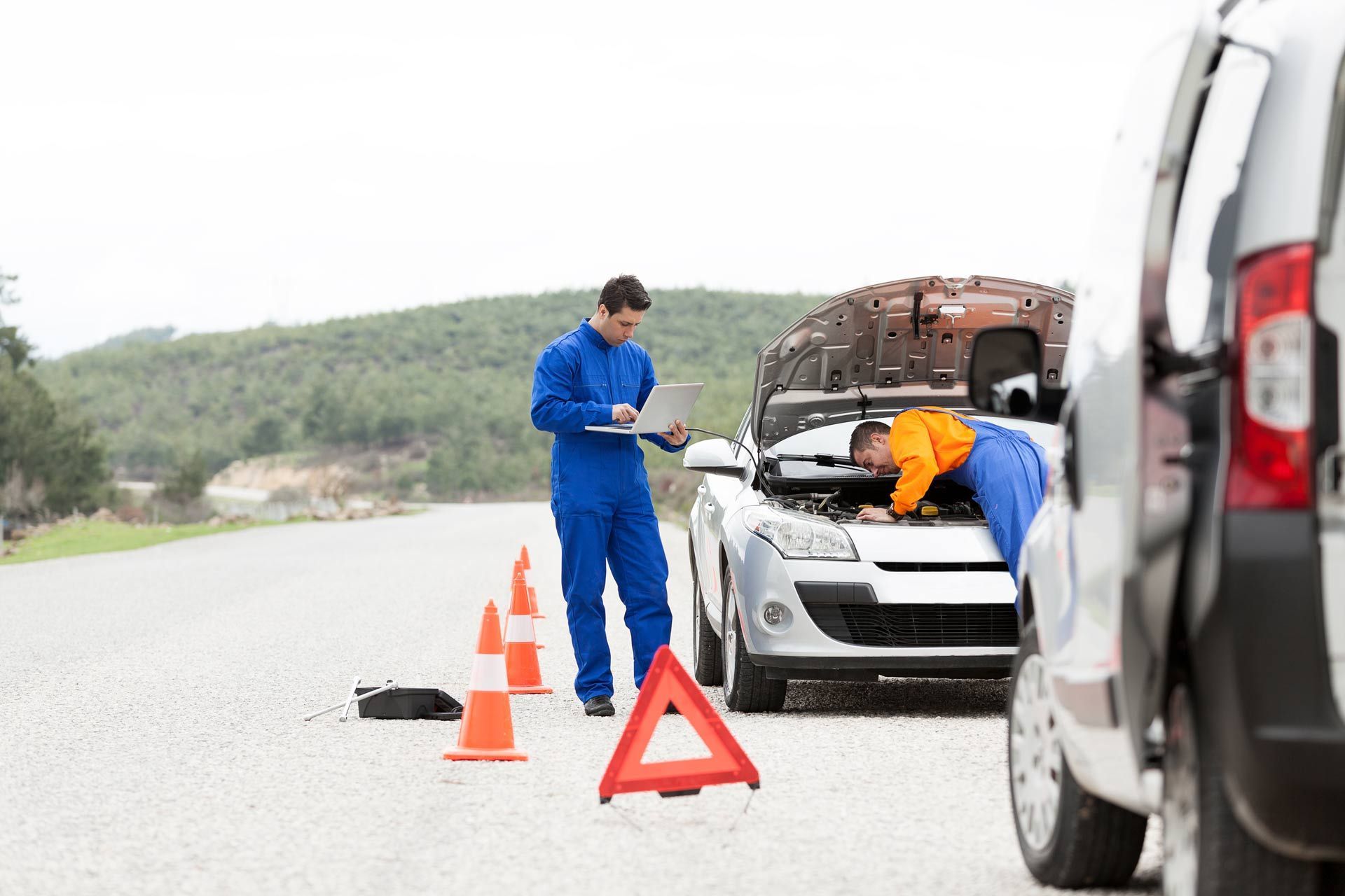 Roadside car breakdown with mechanic checking a car, hazard triangles, and another vehicle nearby on a wet road