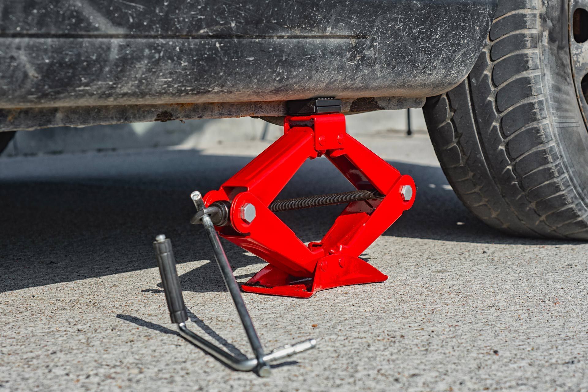 Red car jack lifting a vehicle, with a lug wrench on the pavement beside it.