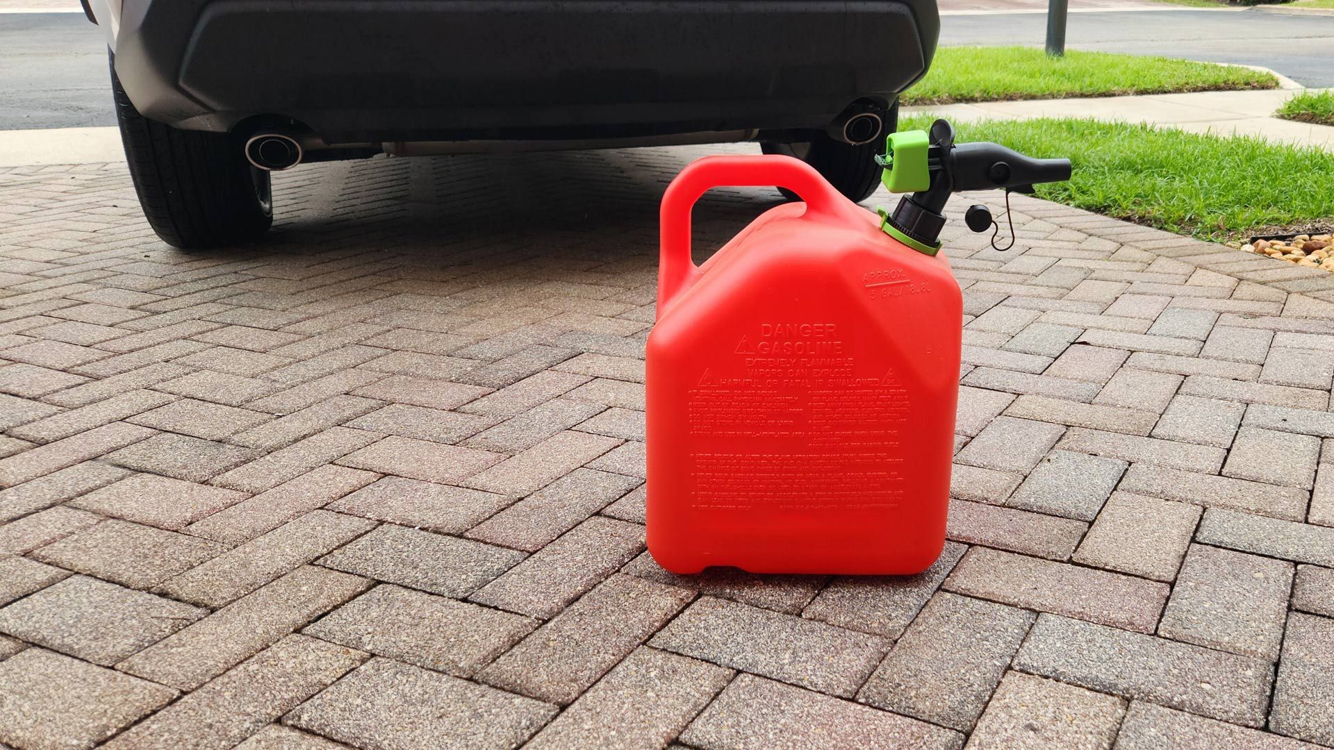 Red gas can on a driveway beneath a car’s rear bumper, with a dangling black hose.