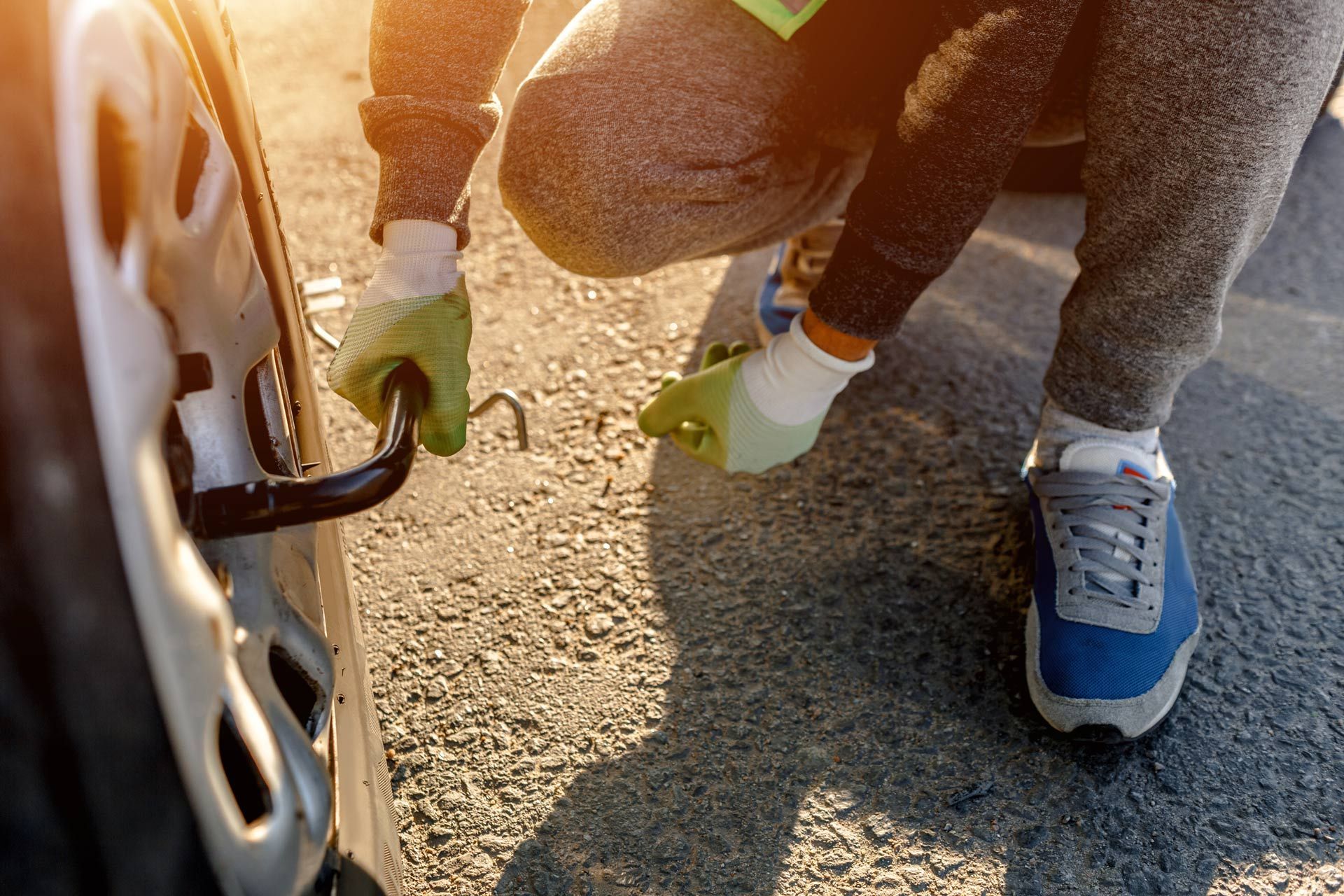 Person kneeling beside a car, using a tool near the wheel on a sunlit gravel road
