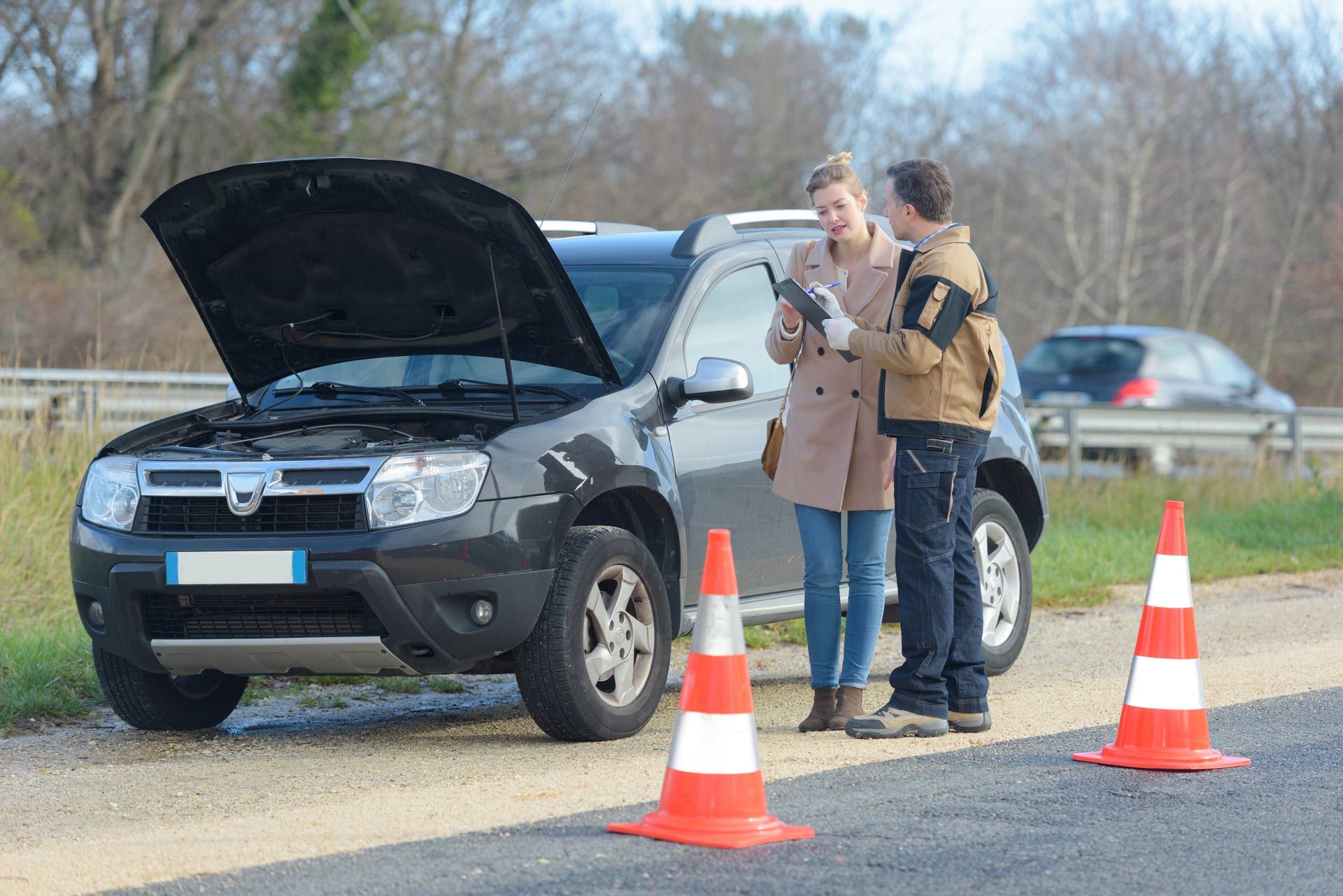 Two people inspect a black SUV with its hood open beside orange traffic cones on a roadside.
