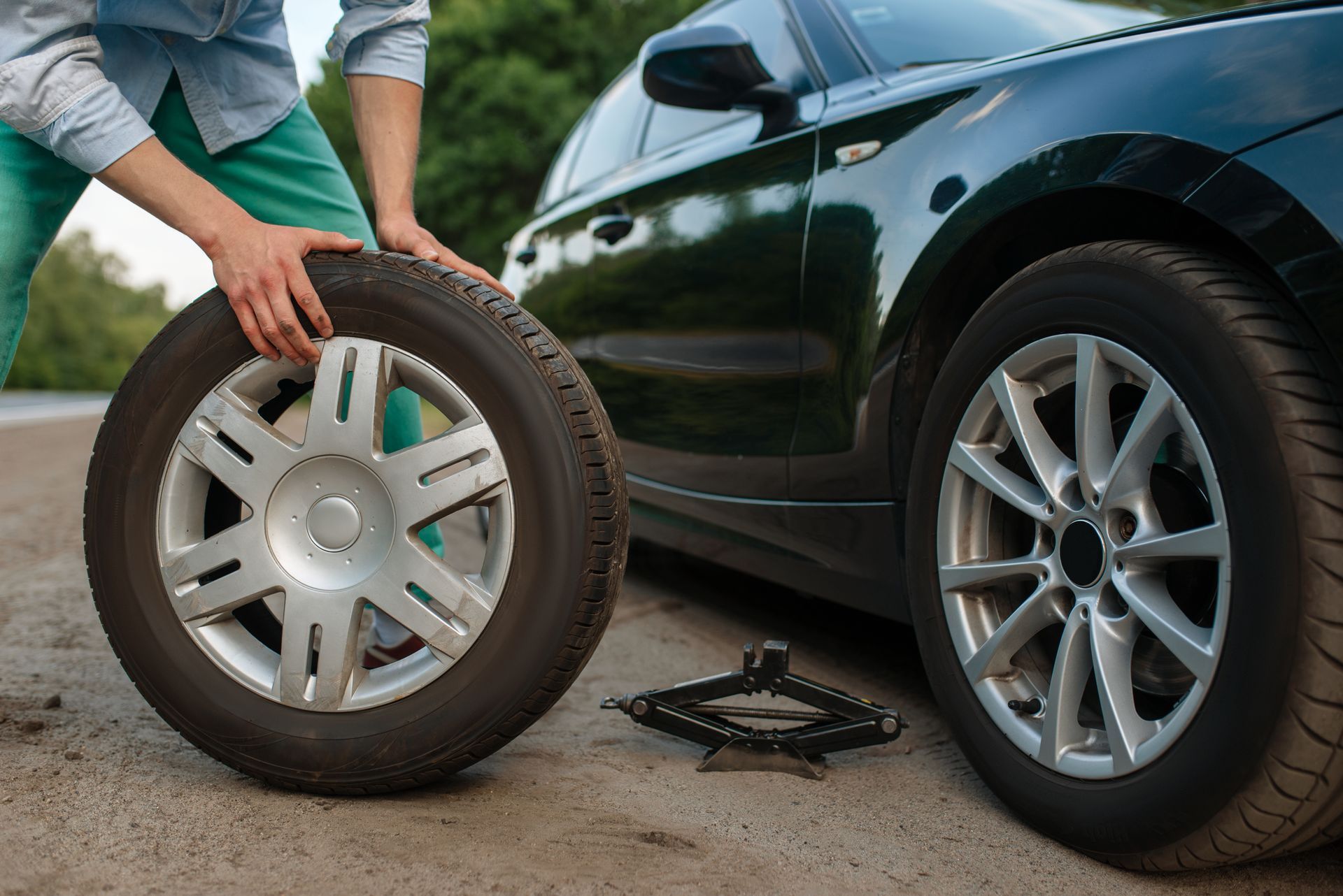 Person changing a car tire beside a black sedan, with a spare wheel and jack on the ground