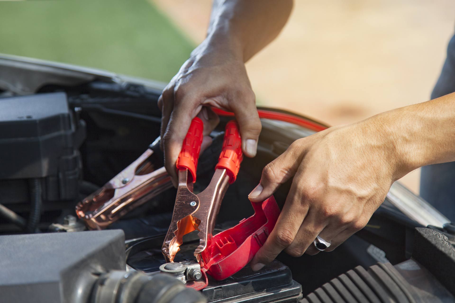Hands connecting red jumper cables to a car battery in an engine bay
