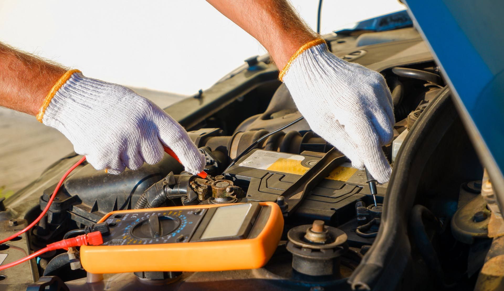 Mechanic’s gloved hands testing a car battery with a multimeter under an open hood
