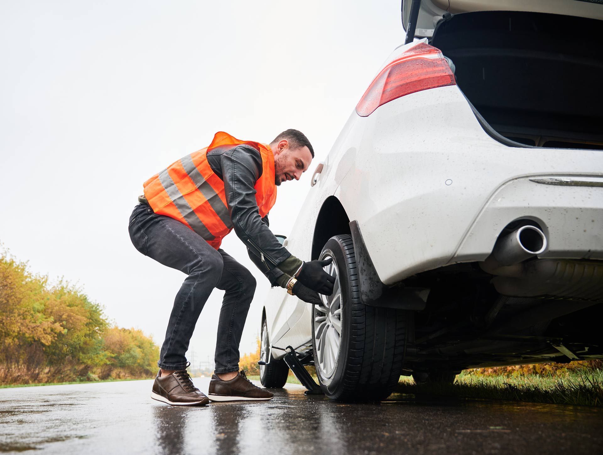 Person in an orange safety vest changing a tire on a white car by the roadside.