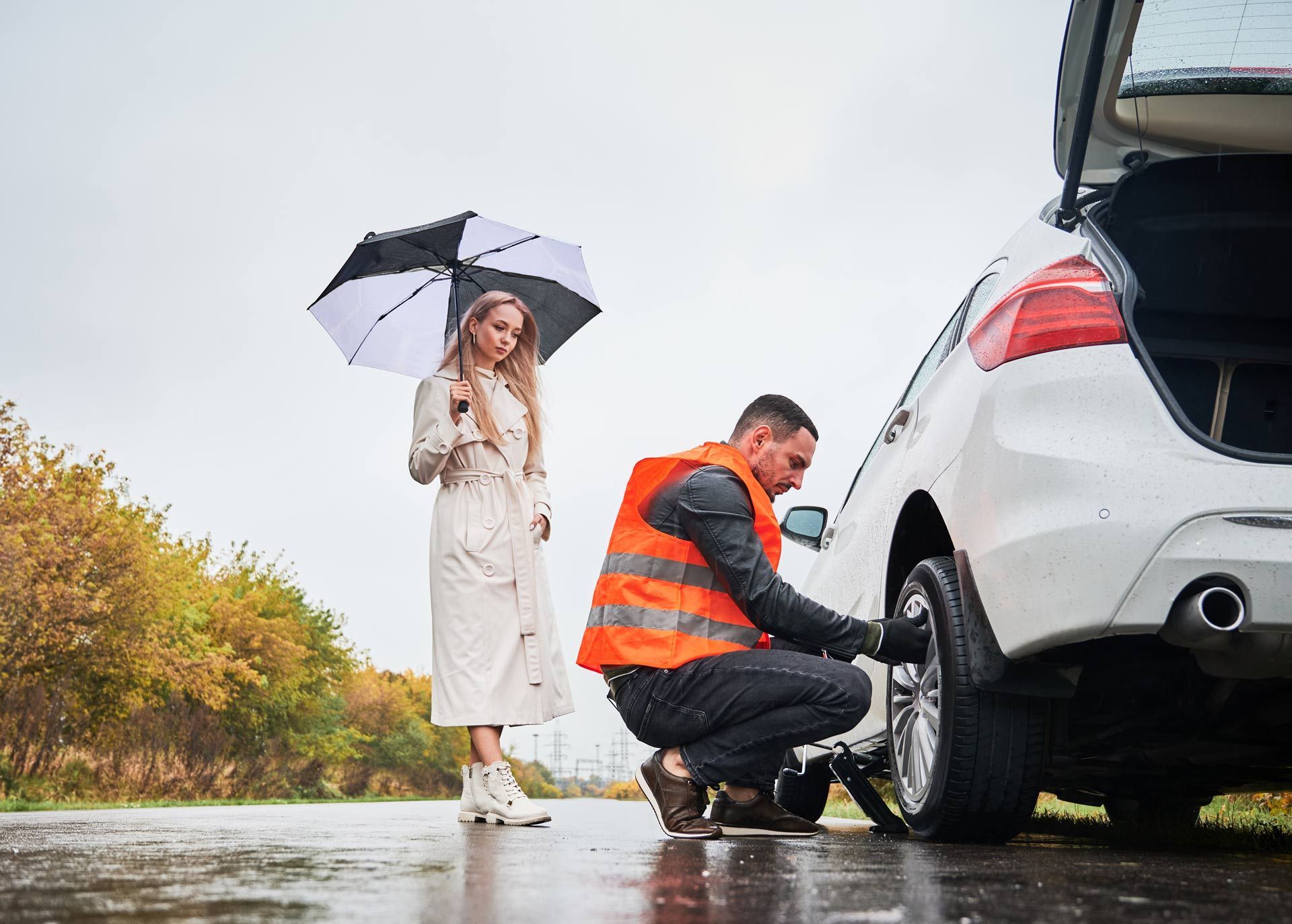 Person changing a car tire in the rain while another holds an umbrella nearby