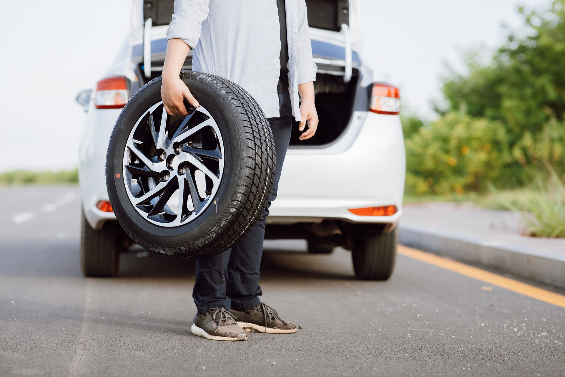 Person holding a spare tire beside a white car on a road.