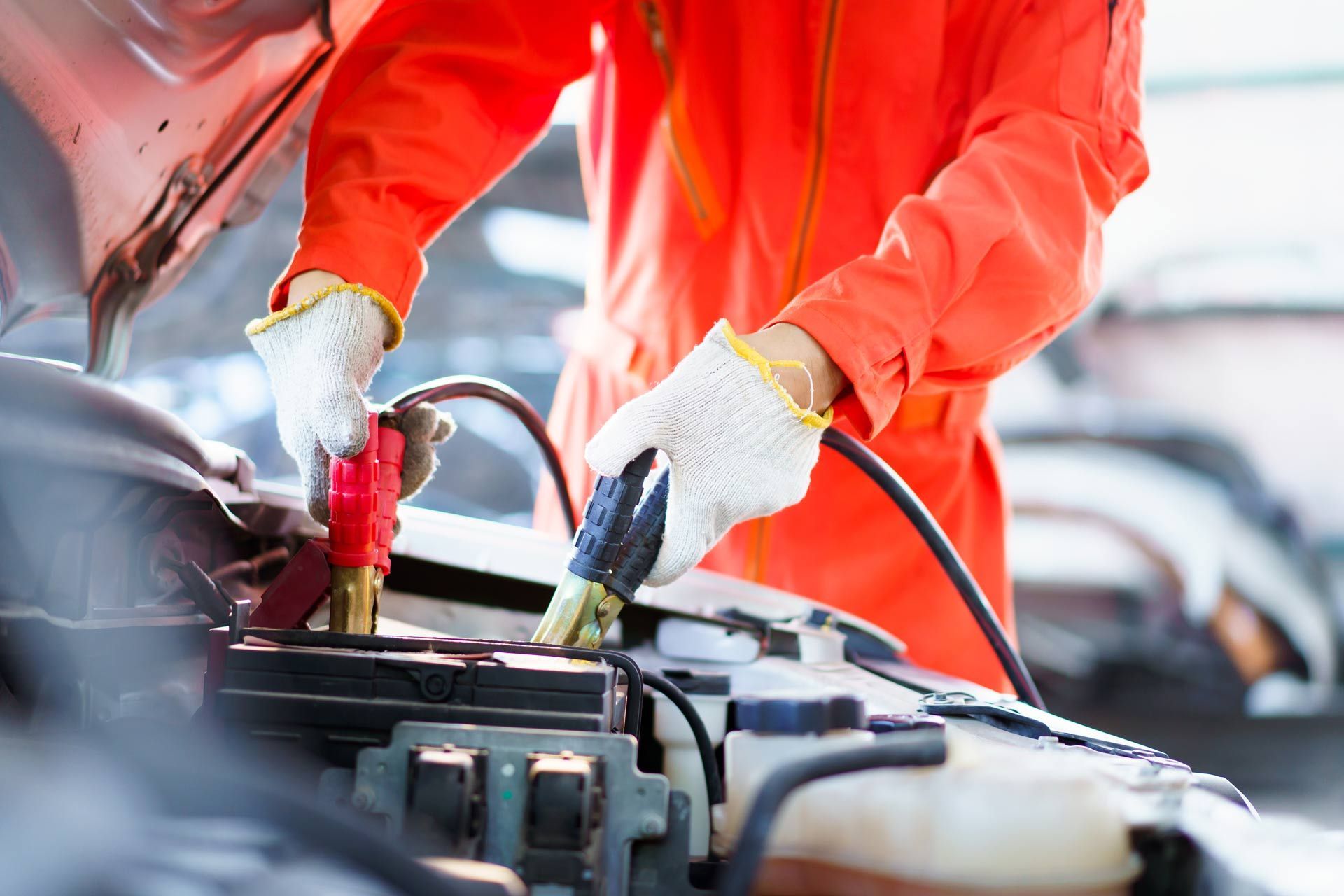 Mechanic in orange jacket using jumper cables to jump-start a car engine in an auto shop