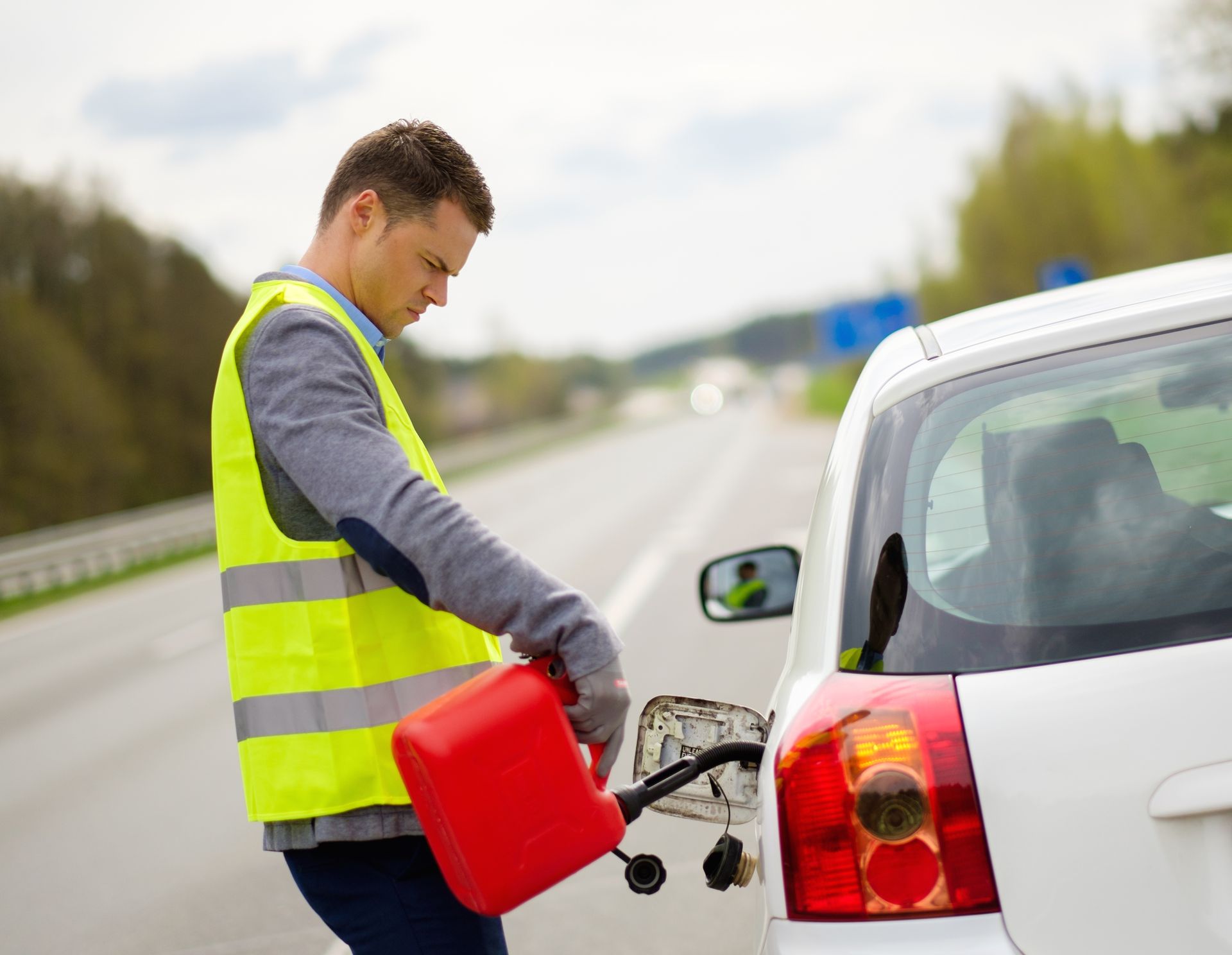 Man in a reflective vest refueling a white car from a red gas can on the roadside