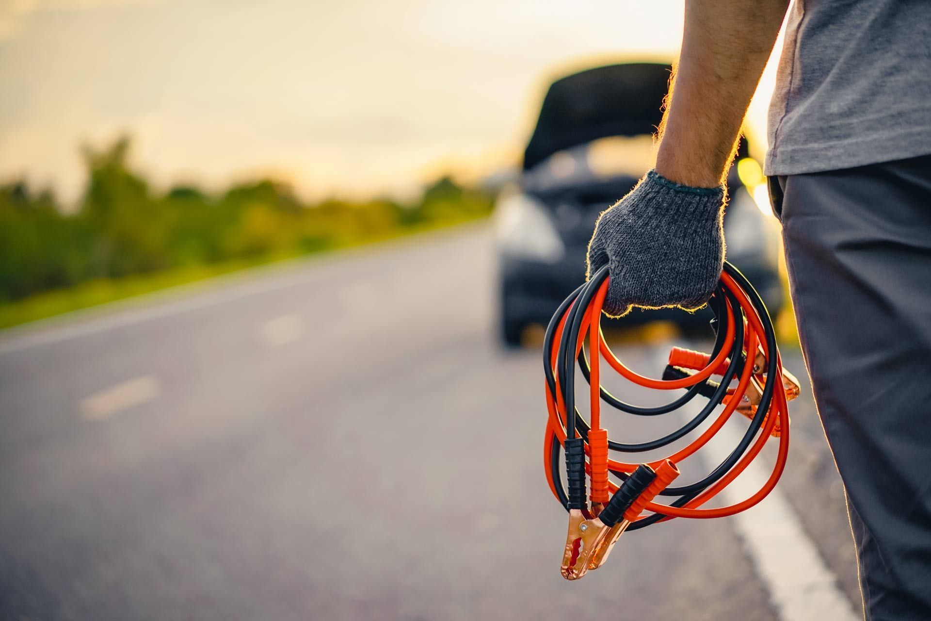 Gloved person holding orange jumper cables beside a roadside car on a sunny day