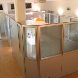Interno di un ufficio con postazioni a cubicolo, pareti divisorie beige e pannelli in vetro satinato sotto un'illuminazione calda.