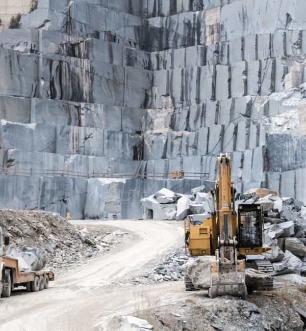 A yellow excavator is driving down a dirt road in front of a large rock wall.