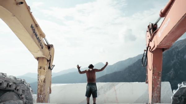 A shirtless man is standing in front of a large excavator.