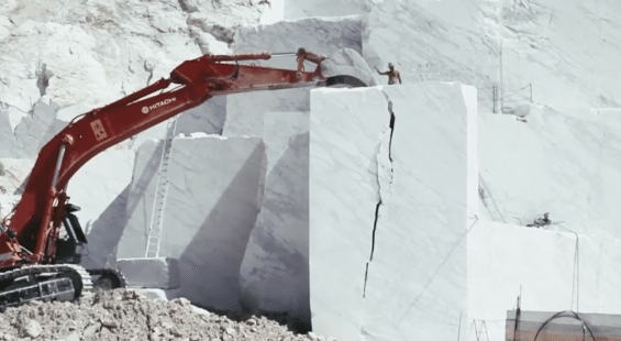 A red excavator is moving a large white block in a quarry.