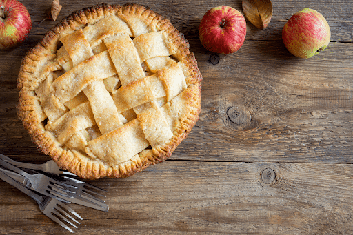 An apple pie is sitting on a wooden table next to apples and forks.