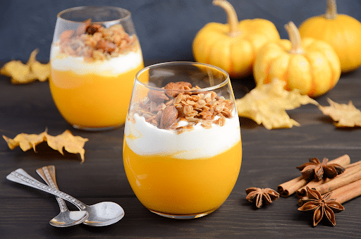A close up of a dessert in a glass on a table with pumpkins in the background.