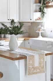 A kitchen with a table runner and a vase of plants on the counter.