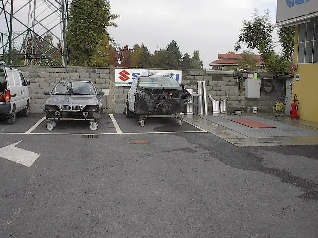 Due auto sverniciate su carrelli parcheggiate vicino a un cartello Suzuki e a un muro di mattoni. Un furgone è parcheggiato sulla sinistra.
