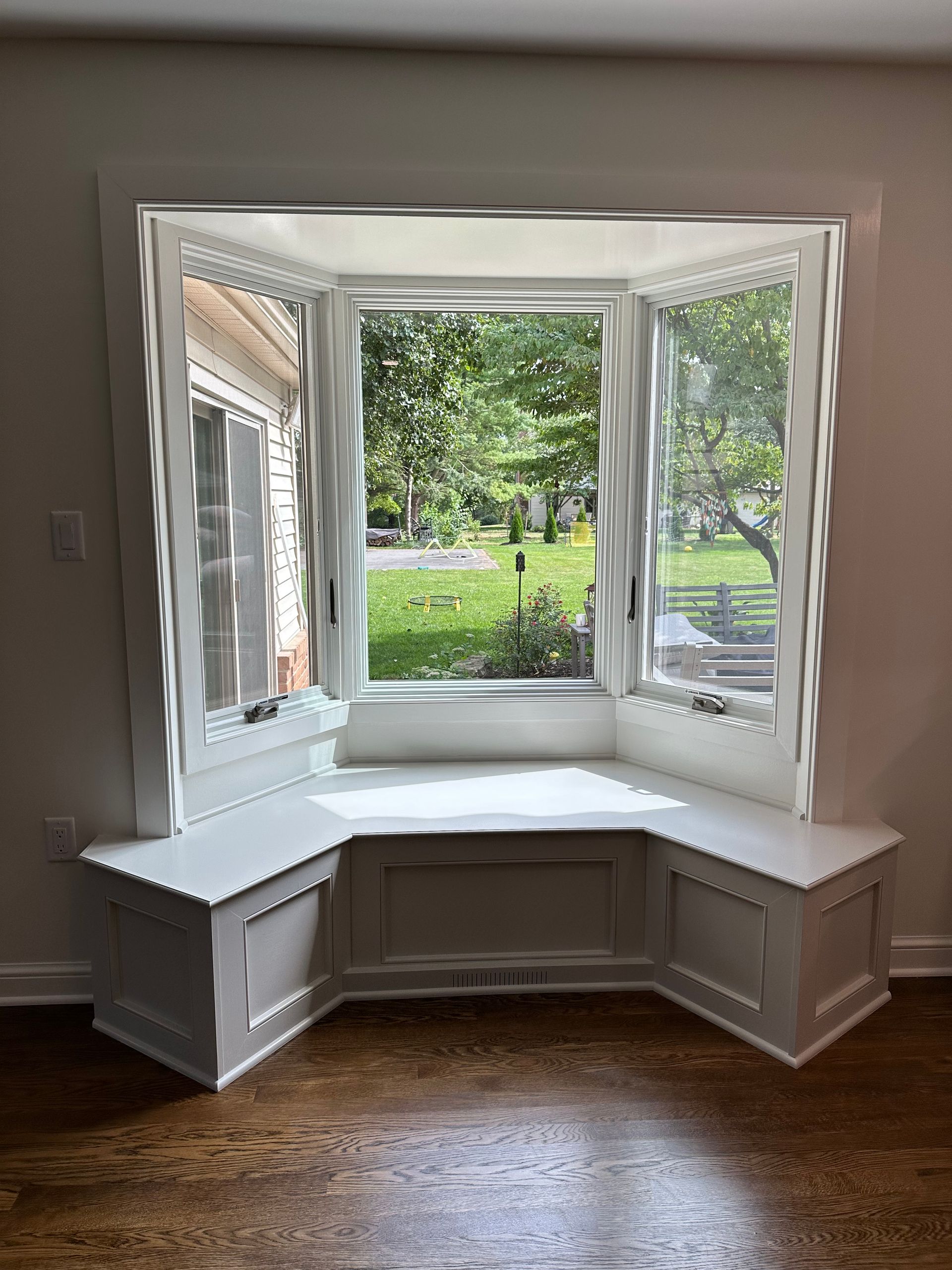 Bay window with built-in seating, painted white, with a view of a green yard and trees.