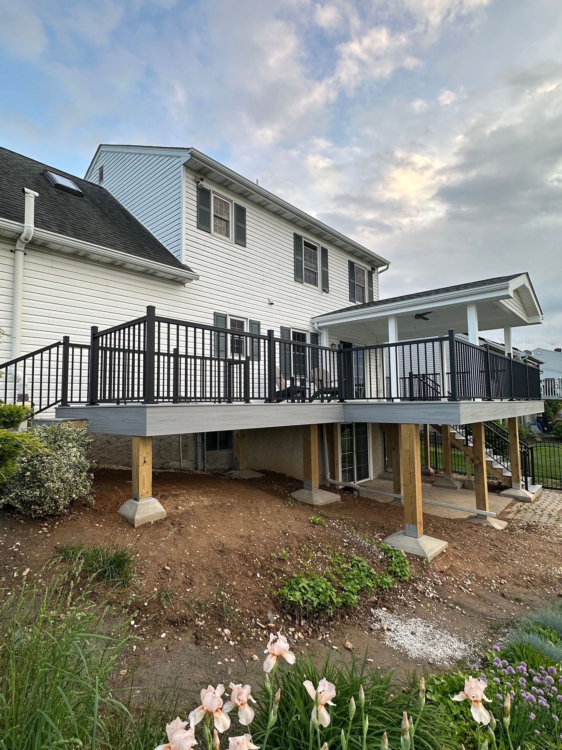 Back of a two-story white house with a gray deck. Black railing surrounds the deck, supported by wooden posts. Cloudy sky.